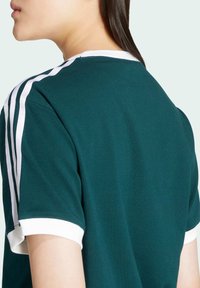 Short-sleeve green t-shirt made of cotton, featuring white accents on the collar and sleeves, with three white stripes on the shoulder.