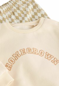 Jasnobeżowy sweatshirt z podniesionymi, jasnobrązowymi literami "HOMEGROWN". Poniżej, wzorzyste spodnie dresowe z naprzemiennie ułożonymi beżowymi i białymi diamentami.