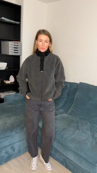 Hall halli flanellist ülerõivas musta kraega, neljandiku lukuga ja logoga. Kandnud tumedate hallide pükstega ja valgete tossudega, istudes türkiissinisel diivanil.