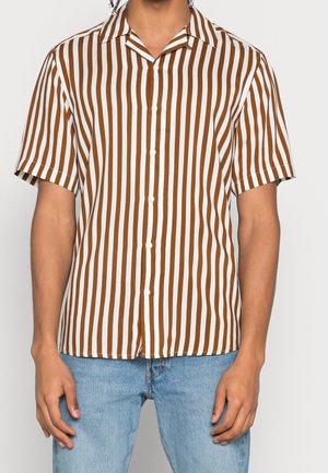 Mann trägt kurzärmliges Hemd mit Kragen mit vertikalen braunen und weißen Streifen und hellblauen Jeans, steht mit entspannten Armen.