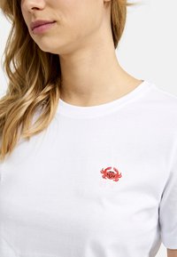Weißes Baumwoll-T-Shirt mit einem roten gestickten Krabbendesign und dem Text "SEA YA" darunter. Runder Halsausschnitt und kurze Ärmel.