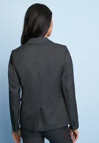 Šedý blazer s klopovým límcem, přiléhavý design, texturovaná tkanina a knoflíkové detaily na rukávech. Ušitý střih s jedním větracím otvorem vzadu.