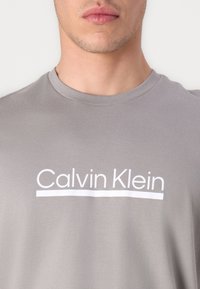 Camiseta de algodón gris con cuello redondo y logo blanco de "Calvin Klein" en el pecho. Textura suave, corte casual.