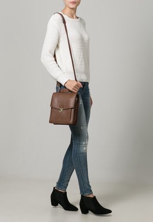 Vrouw draagt een wit gebreide trui, gescheurde skinny jeans, zwarte enkellaarsjes en draagt een bruine leren schoudertas.