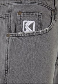 Shorts de mezclilla gris con un borde de bolsillo deshilachado y un parche de logo blanco bordado. El diseño se acentúa con herrajes metálicos.