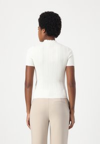 White ribbed short-sleeve top with a high neckline and a fitted design, paired with beige high-waisted trousers, featuring a subtle back detail.