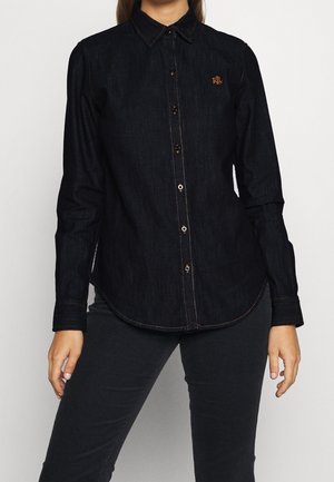 Kvinde iført en tætsiddende sort denim skjorte med knapper og lange ærmer med broderet RLL-logo på brystet, kombineret med sorte bukser.