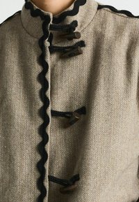 Beige herringbone jacket featuring a high collar, black scalloped trim, and brown toggle fasteners with loops for closure.