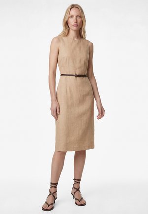 Femme portant une robe beige sans manches jusqu'aux genoux avec une fine ceinture sombre et des sandales à talons marron à brides, debout devant un fond blanc.