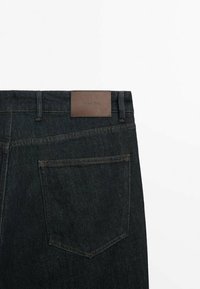 Mörka denimjeans med en strukturerad yta, som har en brun läderlapp vid midjan och en enda bakficka med en spetsig form.