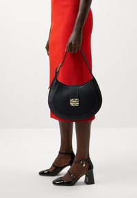 Černá kožená kabelka s zakřiveným tvarem, se zlatým kováním a světlým prošíváním, doplněná černými lakovanými podpatky a červenými šaty.