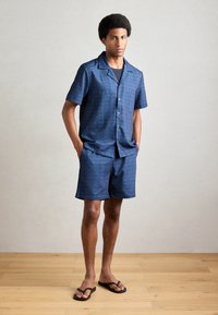 Marineblaue Kurzarmbluse und passende Shorts mit einem sich wiederholenden geometrischen Muster. Leichter Stoff und flache Knöpfe. Getragen mit Flip-Flops.