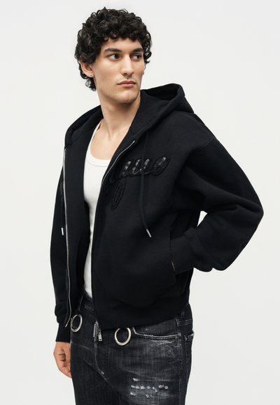 Νέος άντρας με κατσαρά μαλλιά, φορώντας μαύρο φούτερ με φερμουάρ και σκισμένο μαύρο τζιν, κοιτάζοντας προς τα αριστερά με απλό φόντο.