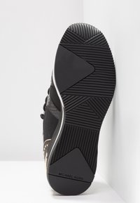 Semelle de basket noire avec motifs de grip texturés et logo "Michael Kors" près du talon, vue par le dessous sur une surface blanche.
