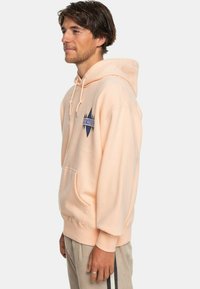 Sudadera de algodón color melocotón con un bolsillo canguro en la parte frontal, capucha con cordón y detalle de logo geométrico azul en el pecho. Se muestra vista lateral.