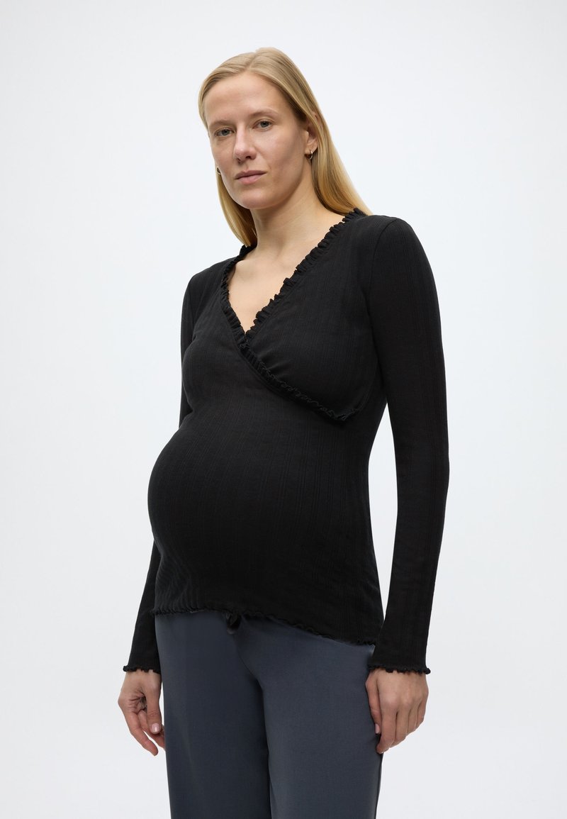 Έγκυος γυναίκα που φοράει μαύρο μακρυμάνικο τοπ τύπου wrap και γκρι παντελόνι, στέκεται μπροστά σε ένα απλό φωτεινό φόντο, κοιτάζοντας την κάμερα.