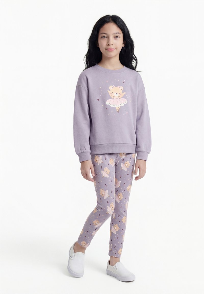 Mädchen trägt einen lavendelfarbenen Pullover und passende Leggings mit Ballerina-Bären-Motiven, steht mit einem Bein überkreuzt, weiße Slipper.