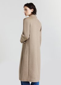 Cappotto in misto lana beige con colletto alto, maniche lunghe e spacchi laterali; lunghezza media e finitura testurizzata. Modello mostrato di schiena.
