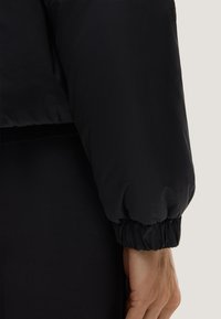 Schwarze Pufferjacke mit einem verkürzten Design. Verfügt über eine glänzende Oberfläche, elastische Bündchen und eine glatte Textur. Minimalistischer Stil ohne Verzierungen.