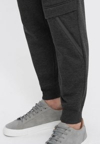Donkergrijze joggingbroek met ritszakken en geribbelde boorden, gecombineerd met lichtgrijze sneakers met een suede textuur en witte zolen.