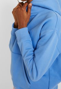Jasnoniebieska bluza z kapturem, wykonana z miękkiego materiału, z kieszenią kangurką i ściągaczami przy mankietach. Kaptur swobodnie opada na szyję.