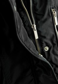 Intérieur de veste noire avec des fermetures éclair argentées, une texture lisse et un détail de bouton-pression. Les fermetures éclair sont placées de chaque côté pour faciliter l'accès.