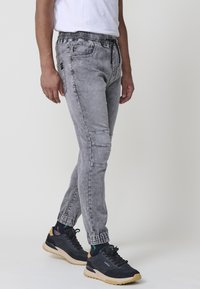 Pantalones jogger grises con cintura elástica y cordón, puños ajustados, bolsillos laterales y un acabado de mezclilla texturizada y desgastada.