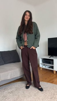 Hosszú sötét hajú nő zöld steppelt kabátot, rövidített rózsaszín pulóvert, barna nadrágot és fekete lapos cipőt visel a nappaliban.