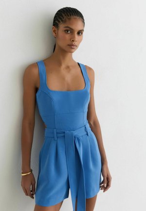 Femme aux cheveux tressés portant une combinaison-short bleue sans manches ceinturée, debout contre un mur blanc texturé.