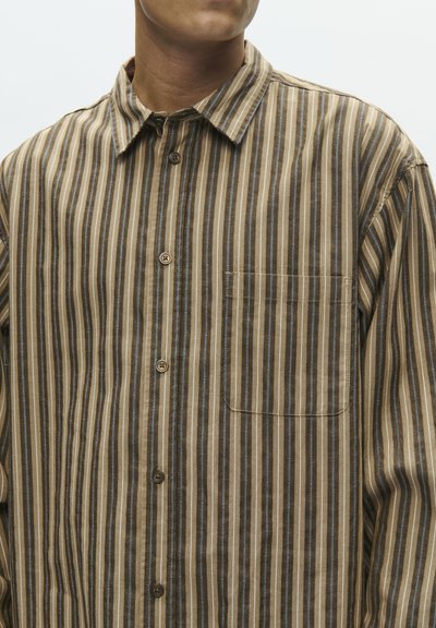 Mężczyzna w brązowej koszuli w paski z guzikami i kieszonką na piersi, pokazujący kołnierzyk i górną część torsu na jednolitym tle.