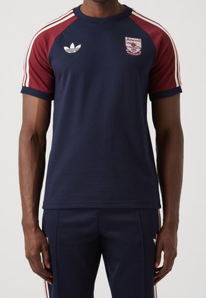 Mężczyzna ubrany w granatowo-bordową sportową koszulkę i spodnie Adidas Arsenal z białymi paskami na rękawach i nogawkach.