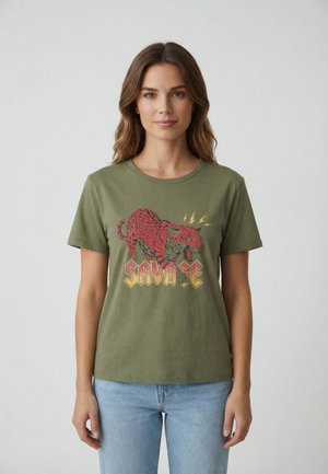 Donna che indossa una t-shirt verde oliva con un leopardo rosso e la parola "SAVAGE" in lettere maiuscole, abbinata a jeans azzurri, in piedi contro uno sfondo semplice.