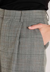 Nahaufnahme einer Hand in der Tasche von grau-braunen Karo-Hosen, die mit einem schwarzen Oberteil getragen werden, wobei die Textur und die Musterdetails des Stoffes zu sehen sind.