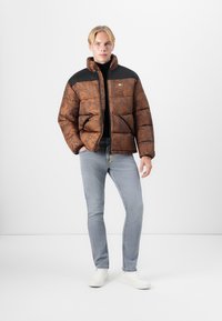 Chaqueta de plumas marrón con un patrón texturizado, hombros negros, bolsillos frontales y un cuello alto, combinada con jeans de un azul claro y zapatillas blancas.
