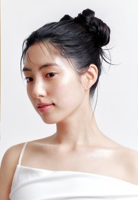 Ritratto di una persona con pelle liscia e chiara e capelli neri acconciati in un morbido chignon, che indossa un top bianco scollato sulle spalle su uno sfondo neutro.