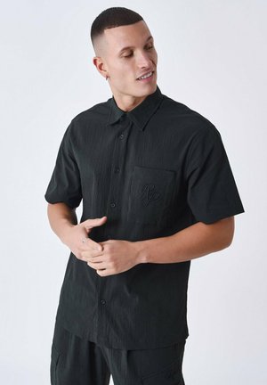 Schwarzes Kurzarmhemd mit strukturiertem Stoff, Knopfleiste, spitzem Kragen und einer Brusttasche mit dezentem Logo-Detail.