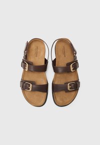 Bruine leren sandalen met twee verstelbare gespsluitingen, een gevoerde suede binnenzool en een platte rubberen zool. Geen teenbedekking, open achterkant ontwerp.