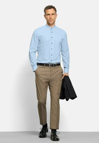 Helles blaues, gepunktetes Hemd mit Knopfverschluss, beige Hose und schwarze Schuhe. Eine dunkle Jacke ist über einen Arm gelegt.