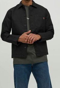 Svart bomullsskjortjacka med avslappnad passform, knäppning, bröstficka och synlig logotyp. Kombinerad med en grön t-shirt och blå jeans.