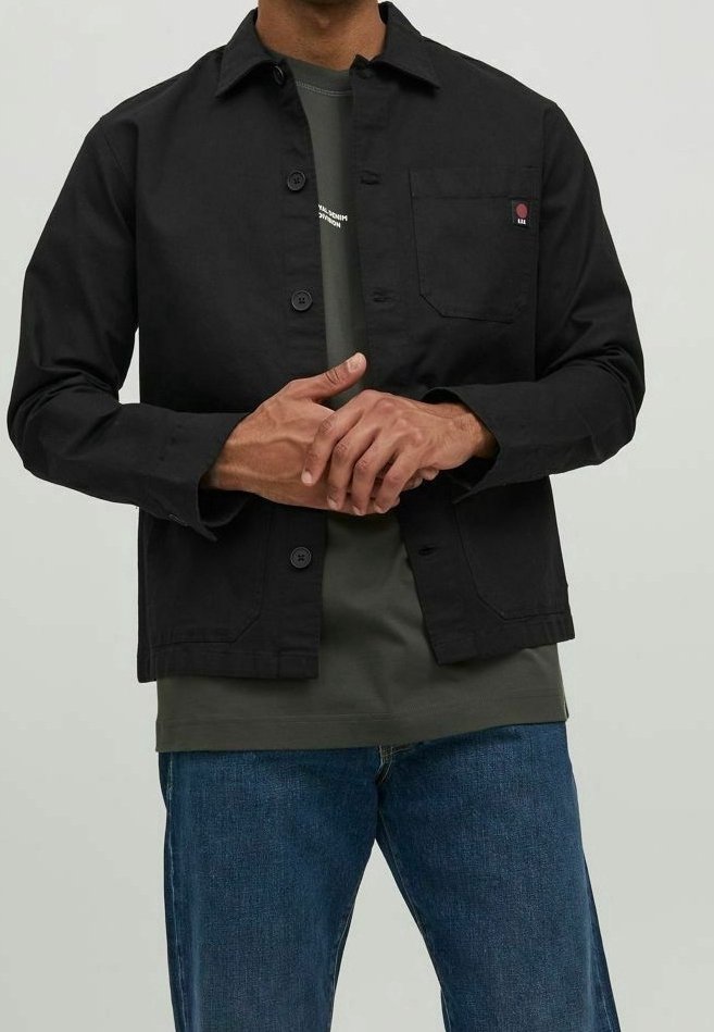 Svart bomullsskjortjacka med avslappnad passform, knäppning, bröstficka och synlig logotyp. Kombinerad med en grön t-shirt och blå jeans.