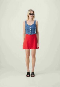 Top in denim senza maniche con chiusura a bottoni, abbinato a una gonna rossa a pois. Il modello indossa sandali neri aperti e occhiali da sole scuri.