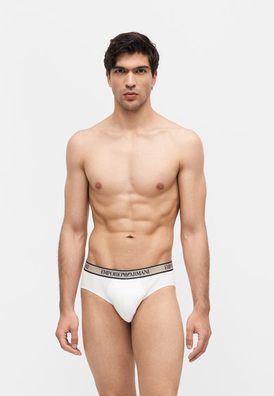 Νέος μυώδης άνδρας στέκεται όρθιος, φοράει λευκά εσώρουχα Emporio Armani με λογότυπο στη μέση, σε απλό ανοιχτό γκρι φόντο.