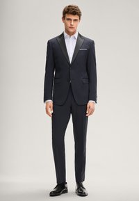 Abito blu navy con risvolti in raso nero, vestibilità sartoriale, design monopetto, abbinato a una camicia bianca e scarpe nere lucide.