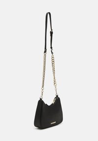 Bolso cruzado de piel negra con forma curva, que presenta un detalle de cadena dorada y asas. Cierre de cremallera y detalle del logo en la parte frontal.