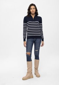 Pull à rayures bleu marine et blanc à manches longues avec zip et poignets côtelés, associé à un jean skinny bleu déchiré et des bottes beiges jusqu'au genou.