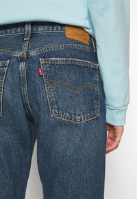 Vaqueros de mezclilla azul con bolsillos traseros cosidos y una etiqueta de cuero marrón de la marca. La textura está ligeramente desgastada con acentos de costura en naranja.