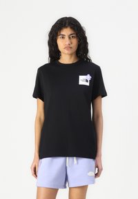 Črna majica s kratkimi rokavi iz bombaža, ki ima belo logotipno nalepko na levem prsnem mestu, dopolnjena z majhnim vijoličnim cvetom.