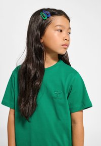 Grön bomullströja med rund hals och korta ärmar, med en broderad logotyp på vänster bröst. Långt mörkt hår med hårklipp.