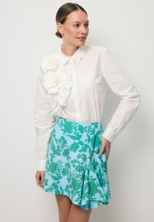 Kobieta ubrana w białą bluzkę z dużymi kwiatami z tkaniny i niebieską spódnicę w zielone wzory kwiatowe, pozująca z jedną ręką na biodrze.