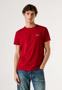 Rood katoenen T-shirt met een ronde hals en korte mouwen, met een klein wit logo op de linkerborst. Gecombineerd met blauwe spijkerbroek.
